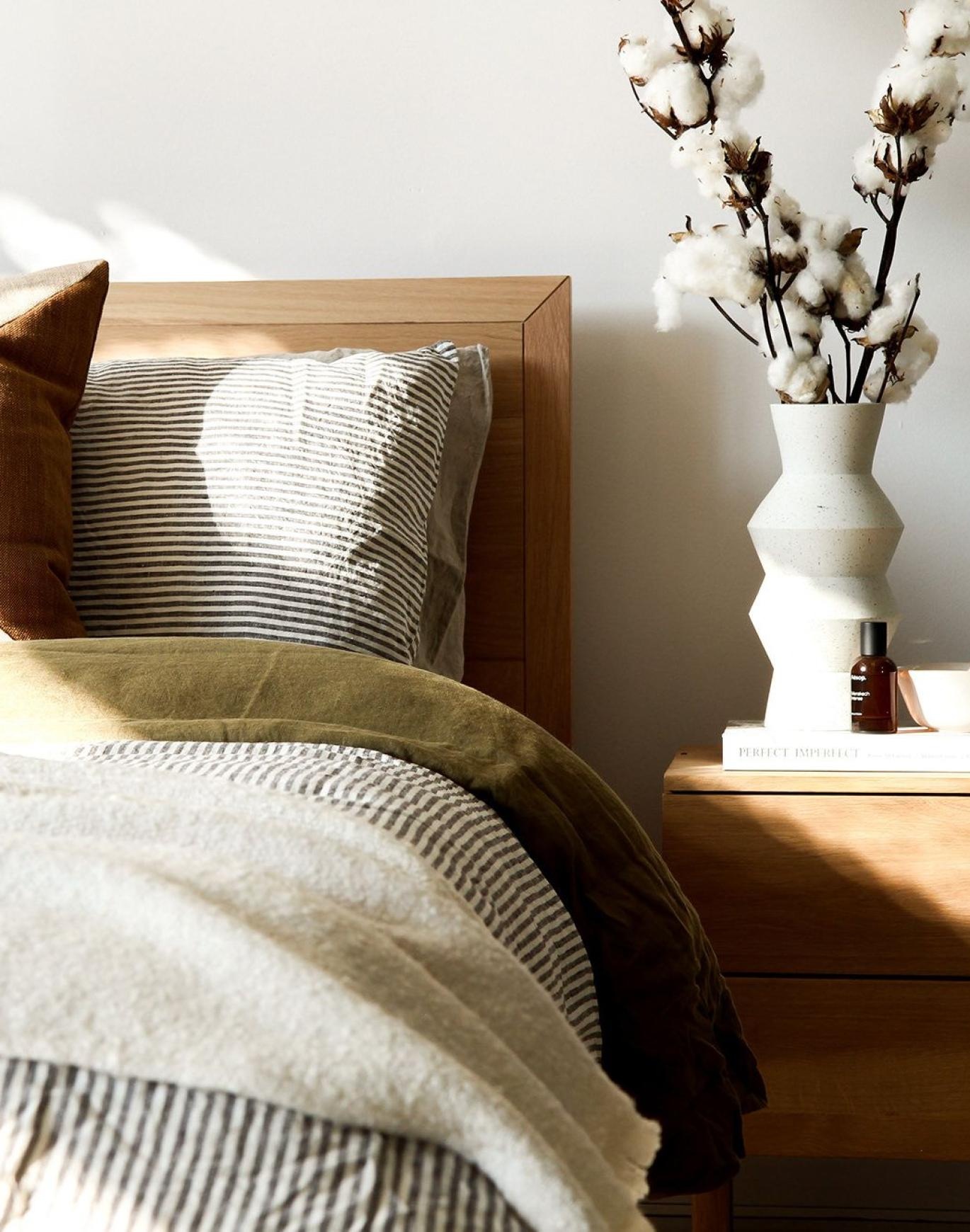 swath of sunlight shines across a well made bed and bedside table with a vase of cotton plants